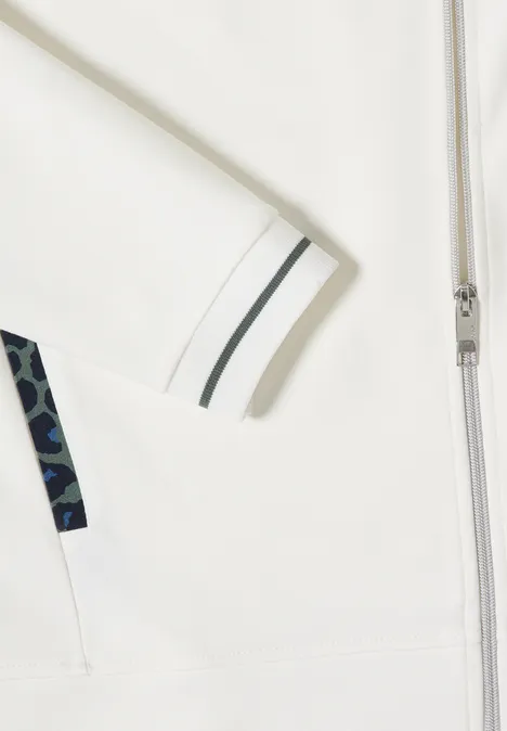 Detail eines weißen Kleidungsstücks mit Reißverschluss, gestreiftem Ärmelbund und geometrischem Muster.