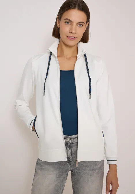 Frau in weißer Kapuzenjacke mit schwarzen Kordeln, blauem Top und Jeans.