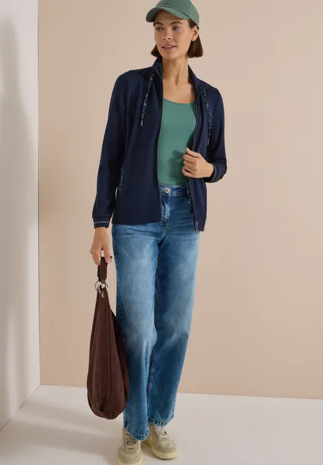 Eine Frau trägt grünes Top, marineblaue Jacke, blaue Jeans, Cap und hält eine braune Tasche.