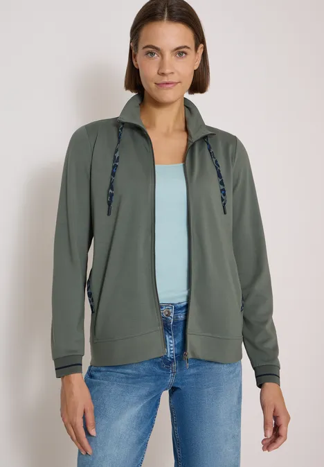 Eine Frau in leichter olivgrüner Kapuzenjacke über hellblauem Top und Jeans steht vor neutralem Hintergrund.