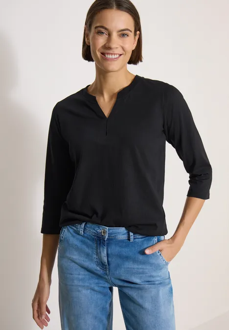 Frau in schwarzem Langarmshirt und blauen Jeans steht vor hellem Hintergrund.