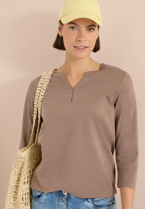 Person trägt beigefarbenes Langarmshirt und geflochtene Tasche vor neutralem Hintergrund.