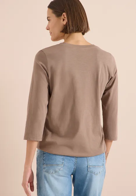 Frau von hinten in taupefarbenem 3/4-Arm-Pullover und blauen Jeans vor neutralem Hintergrund.