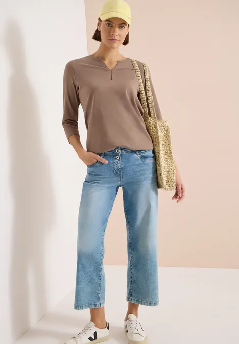 Frau in hellbraunem 3/4-Arm-Shirt, hellblauen Culotte-Jeans, weißen Sneakers und Strohtasche vor neutraler Wand.