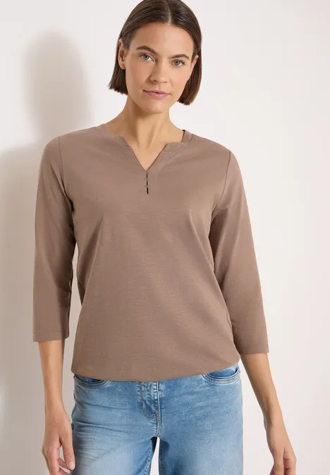 Person trägt ein taupefarbenes 3/4-Shirt mit kurzem Reißverschluss am Kragen und blaue Jeans vor hellem Hintergrund.