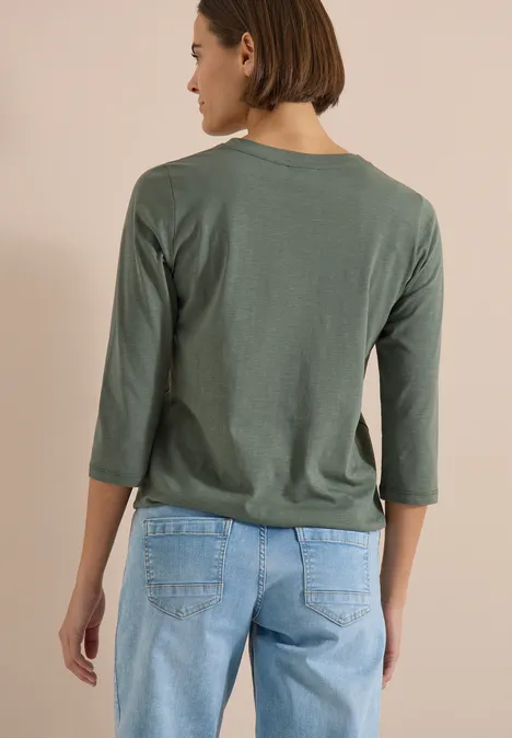 Rückansicht einer Person mit grünem 3/4-Arm-Shirt und blauen Jeans vor beigem Hintergrund