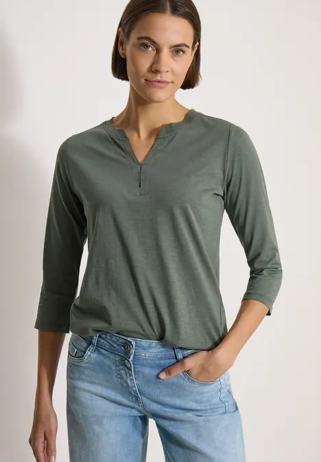 Frau trägt grünes Shirt mit 3/4-Ärmeln und V-Ausschnitt, halb in helle Jeans gesteckt.