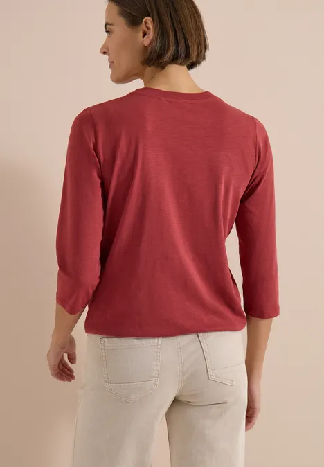Eine Person in rotem Dreiviertelarm-Shirt und heller Jeans, von hinten vor neutralem Hintergrund.