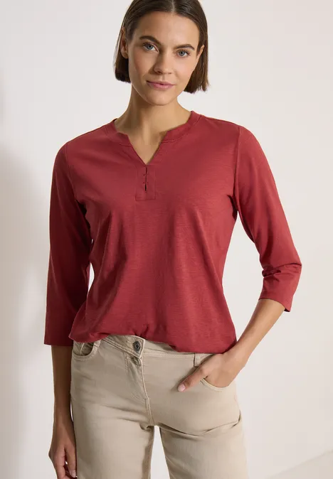 Frau trägt kastiges, dunkelrotes 3/4-Arm-Henley-Shirt mit Knopfleiste und eine beigefarbene Hose.