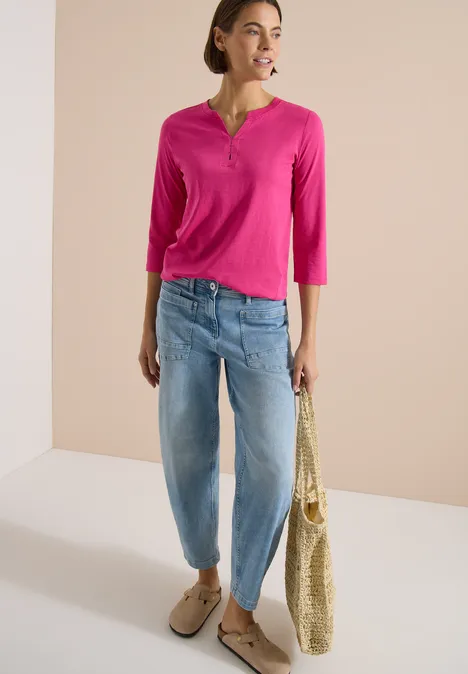 Frau trägt pinkes Henley-Shirt, helle Boyfriend-Jeans, Basttasche und beige Loafers vor hellem Hintergrund.