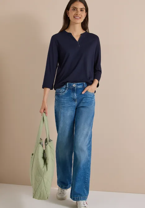 Frau in dunkelblauer Bluse, weiten Jeans und grüner Tasche mit Bommel vor neutralem Hintergrund.
