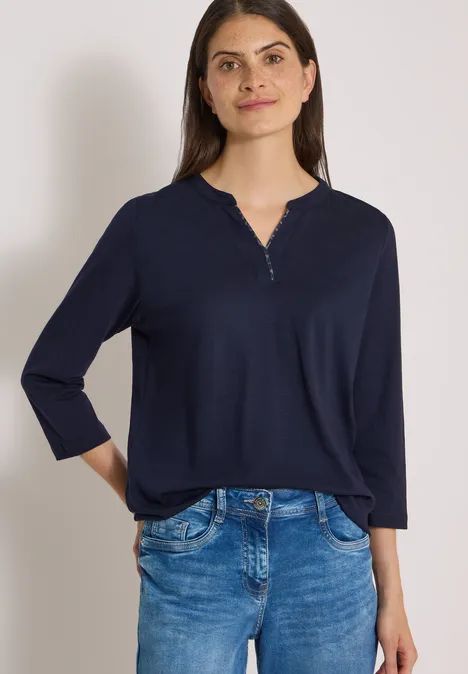 Frau trägt dunkelblaues Shirt mit 3/4-Ärmeln und Jeans vor weißem Hintergrund.