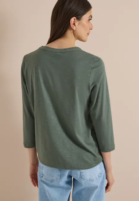 Frau in olivgrünem 3/4-Arm-Shirt und hellblauen Jeans, Rückenansicht vor beigem Hintergrund.