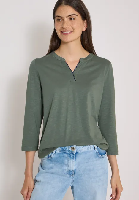 Frau lächelnd in grünem Langarmshirt und blauer Jeans vor weißem Hintergrund