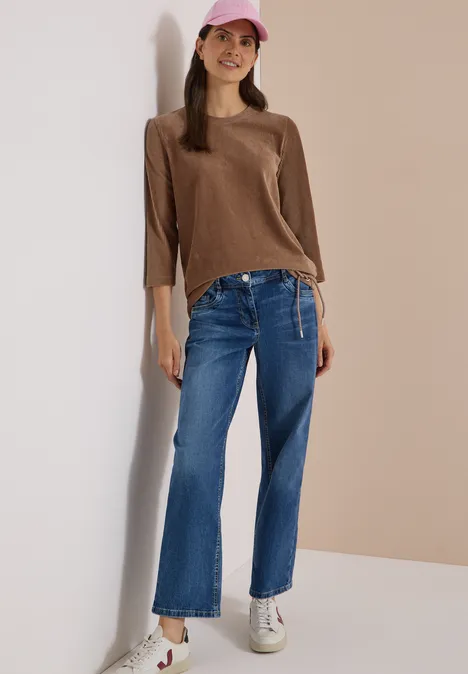 3/4-mouw Shirt van corduroy met trekkoord creamy mocha Eine junge Frau lehnt fröhlich an der Wand, trägt braunen Pullover, weite Jeans, weiße Sneaker und rosa Cap.