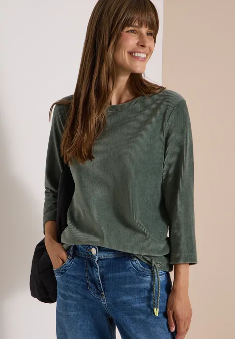 Eine lächelnde Frau in grünem Langarmshirt und blauen Jeans hält eine Hand in der Hosentasche.