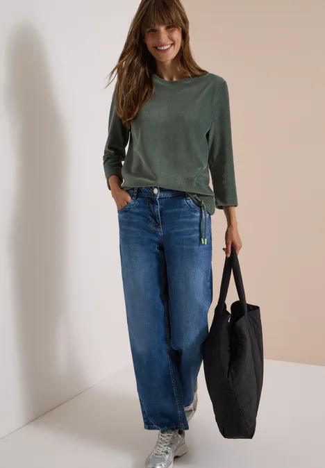 3/4-mouw Shirt van corduroy met trekkoord dried olive green Lächelnde Frau lehnt an Wand, trägt grünes Shirt, weite Jeans und schwarzen Shopper.