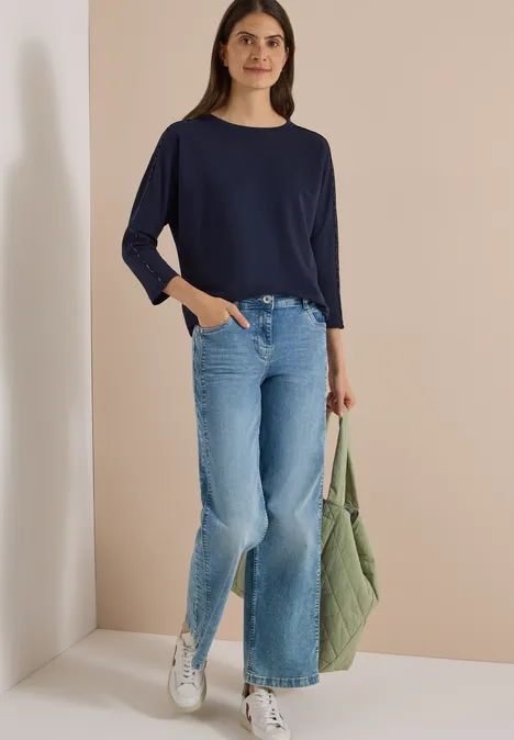 Junge Frau mit dunkelblauem Langarmshirt, hellen Schlagjeans, weißen Sneakern und grüner Tasche vor beigem Hintergrund.