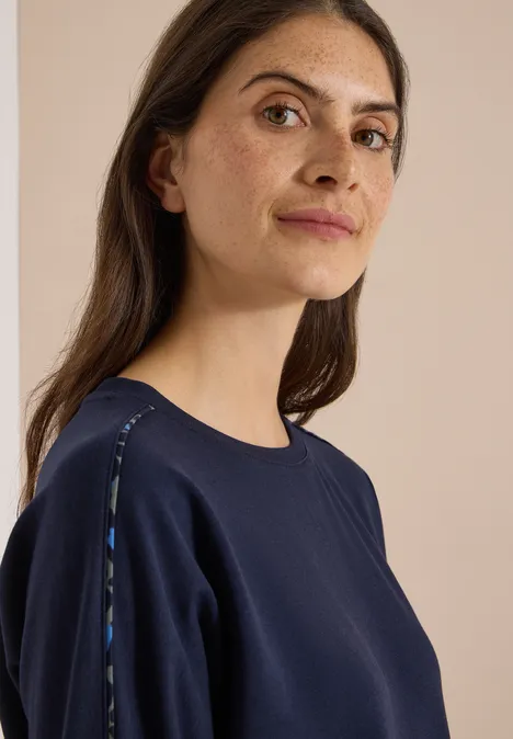 Eine Frau in dunklem Sweatshirt mit dezenten Schulterdetails blickt ruhig zur Kamera.