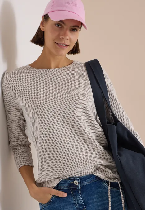 Frau in beiger Longsleeve, Jeans, rosafarbenem Cap und blauer Tasche lehnt an Wand.