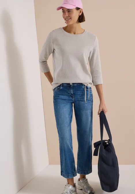 Frau mit rosa Cap, grauem 3/4-Shirt, blauen Cropped-Jeans, weißen Sneakern und navy-Tasche.