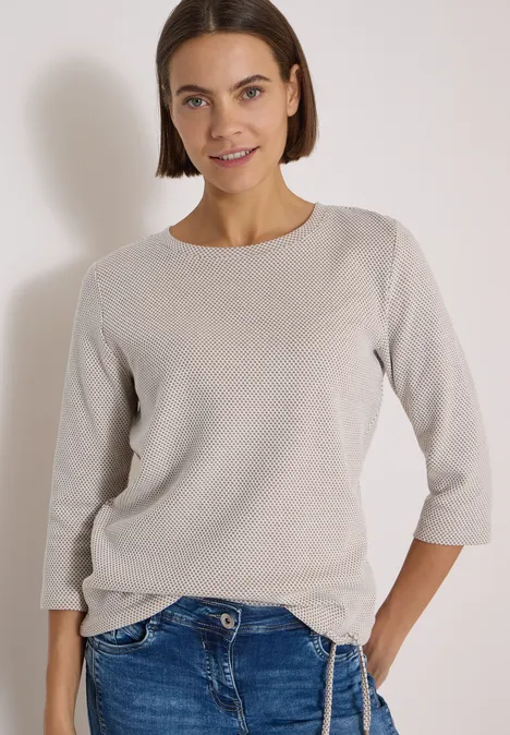 Eine junge Frau mit kurzem Bob trägt einen hellgrauen 3/4-Ärmel-Pullover und Jeans, sie lächelt.