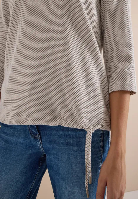 Person trägt beige, grobmaschigen Pullover mit seitlicher Schleife und blaue Jeans.
