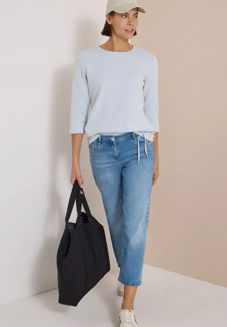 Frau in hellblauem Pullover, Jeans, weißer Cap und großer schwarzer Tasche vor neutralem Hintergrund.