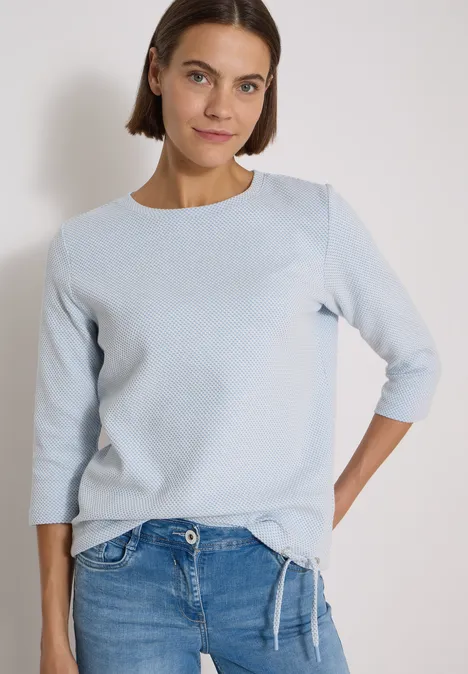 Frau mit schulterlangem Haar in hellblauem Shirt und Jeans vor neutralem Hintergrund.