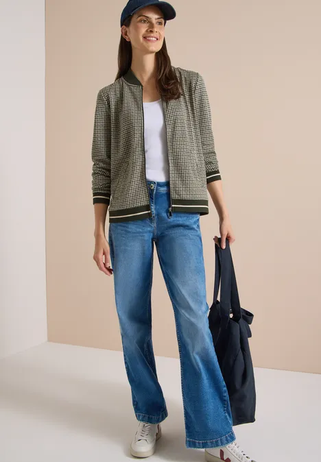 Eine junge Frau in Jeans, weißem Shirt, khakifarbener Jacke, Sneakers und Cap trägt eine große blaue Tasche.