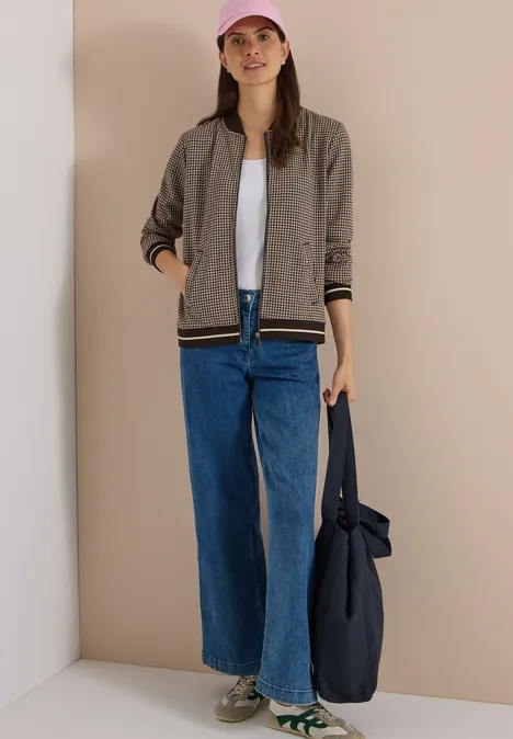 Eine Frau mit rosa Cap, grauer Bomberjacke, weißem Shirt, weiten Jeans hält eine blaue Tasche.