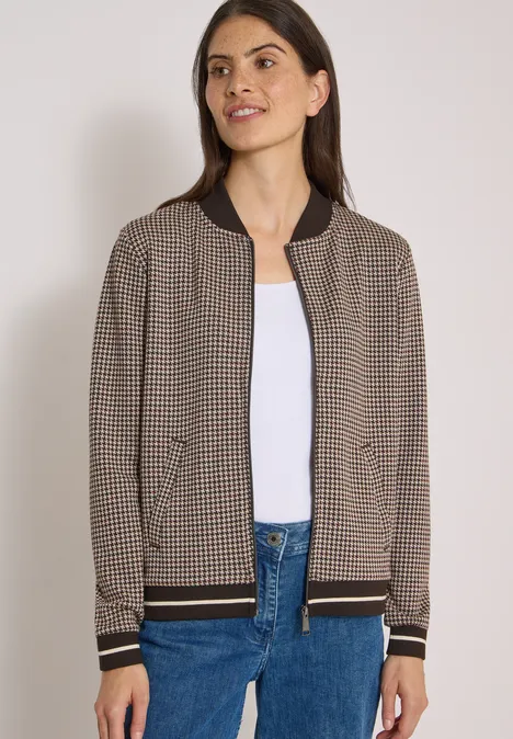 Frau trägt karierte braune Bomberjacke mit schwarzen Bündchen, weißes Shirt und blaue Jeans.