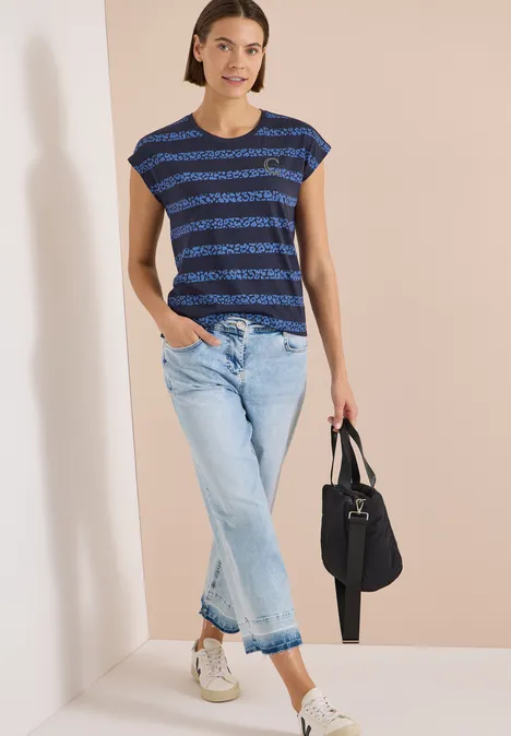 Frau trägt gestreiftes blaues T-Shirt, helle Jeans, weiße Sneaker und hält eine schwarze Tasche.