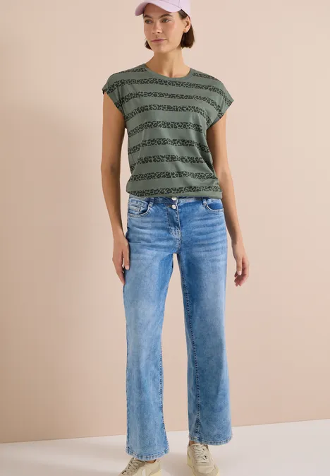 Person trägt weite blaue Jeans und ein gestreiftes grün-schwarzes T-Shirt vor hellem Hintergrund.