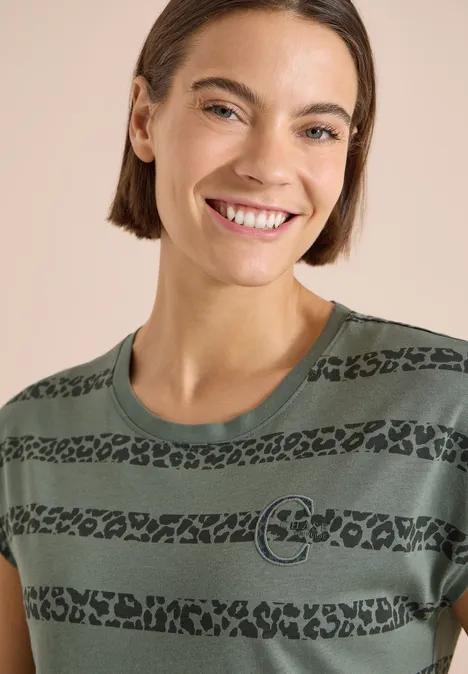 Person mit kurzem Haar trägt grünes T-Shirt mit schwarzen Leopardenstreifen und Kreislogo vor hellem Hintergrund.