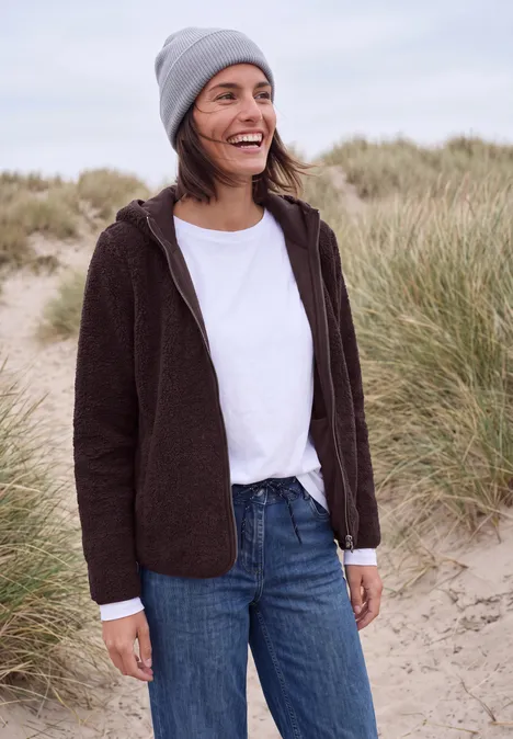 Eine lächelnde Frau mit grauer Mütze, brauner Jacke und Jeans steht am Strand zwischen Dünen.