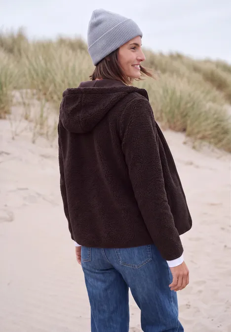 Frau mit brauner Fleecejacke und grauer Mütze steht lächelnd am Strand zwischen Dünen.