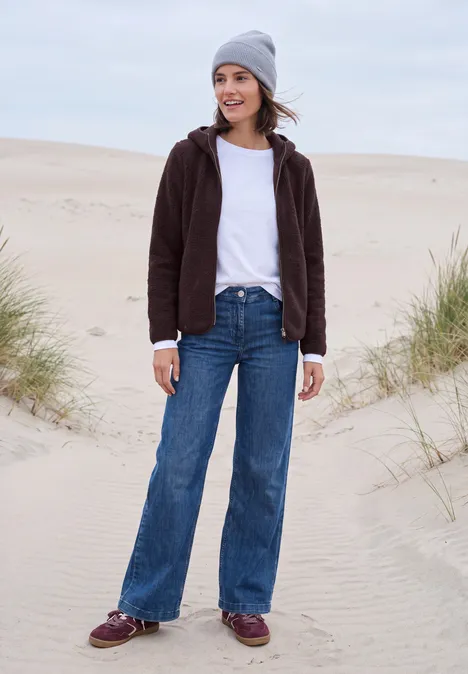 Junge Frau in grauer Mütze, brauner Jacke und weitem Jeans steht lächelnd auf sandigen Dünen.