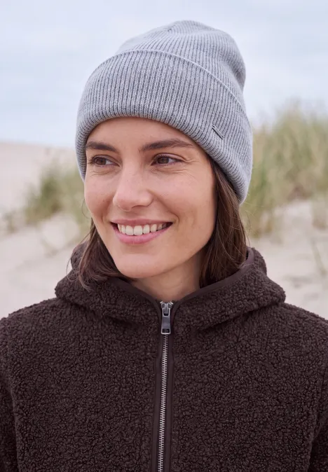 Lächelnde Frau mit grauer Strickmütze und brauner Jacke am Sandstrand im Hintergrund.
