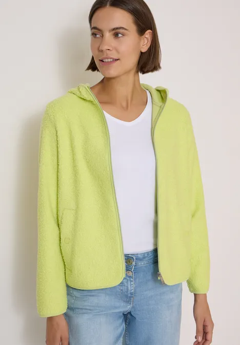Junge Frau in hellgrüner Fleecejacke mit Kapuze über weißem Top und Jeans.