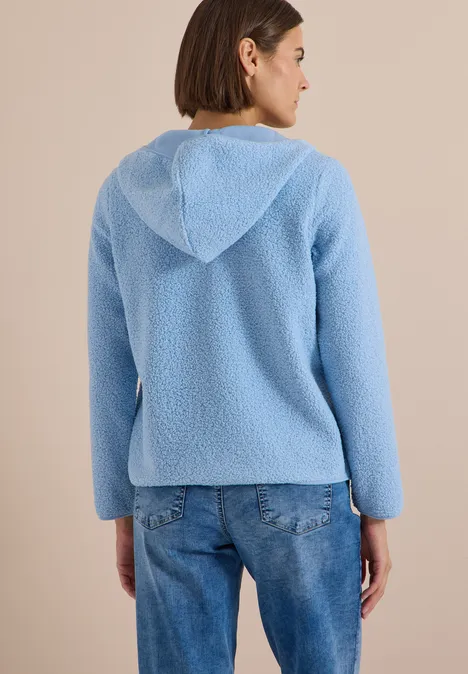 Eine Frau von hinten in einer hellblauen, flauschigen Kapuzenjacke und Jeans vor beigem Hintergrund.