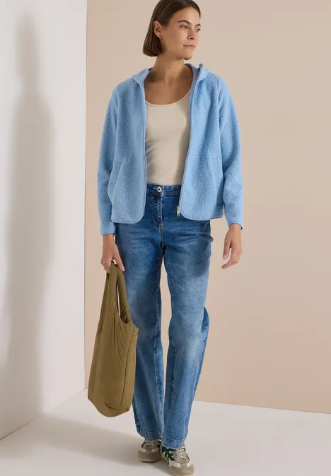 Frau trägt blaue Fleecejacke, weißes Top, Jeans und trägt grüne Tasche