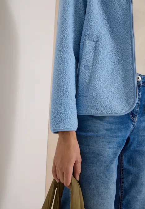 Teilansicht einer Person in Jeans, flauschiger hellblauer Jacke und grüner Tasche.