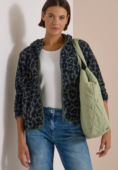 Frau in Jeans, weißem Top und gemusterter Jacke trägt eine grüne gesteppte Tasche über der Schulter.