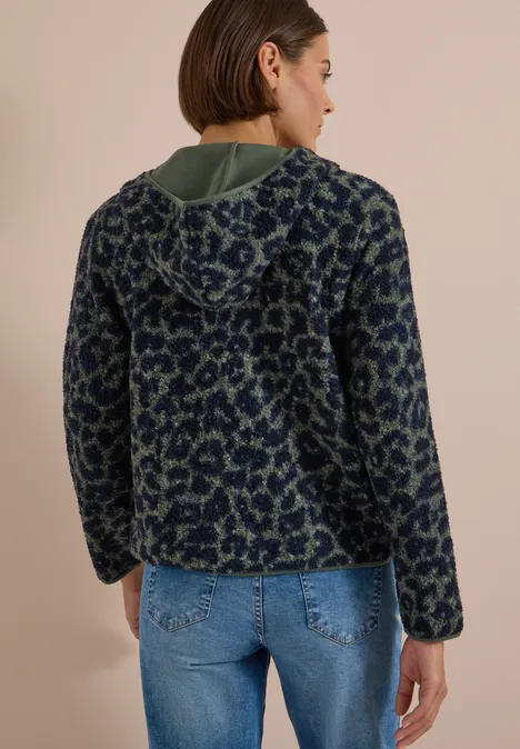 Rückansicht einer Frau in einer blau-grünen gemusterten Fleecejacke mit Kapuze und Jeans vor beigem Hintergrund.