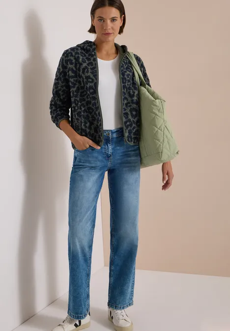 Frau in weißem T-Shirt, blauen Jeans, gefleckter Fleecejacke, mit gesteppter Tasche und weißen Sneakern.