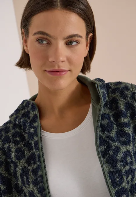 Eine Frau mit kurzem braunem Haar trägt eine grüne Fleecejacke mit Animal-Print und weißes Shirt.