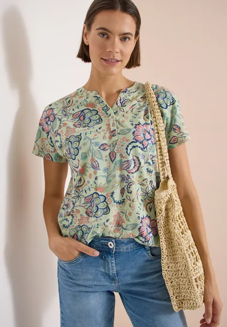 Frau trägt geblümtes Kurzarm-Shirt, blaue Jeans und gestaltet mit Strohtasche.