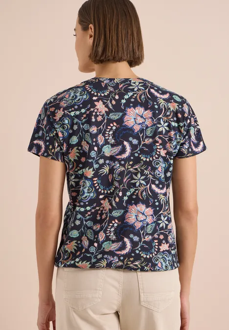 Frau von hinten in dunkelblauem T-Shirt mit buntem Blumenmuster und hellen Hosen.