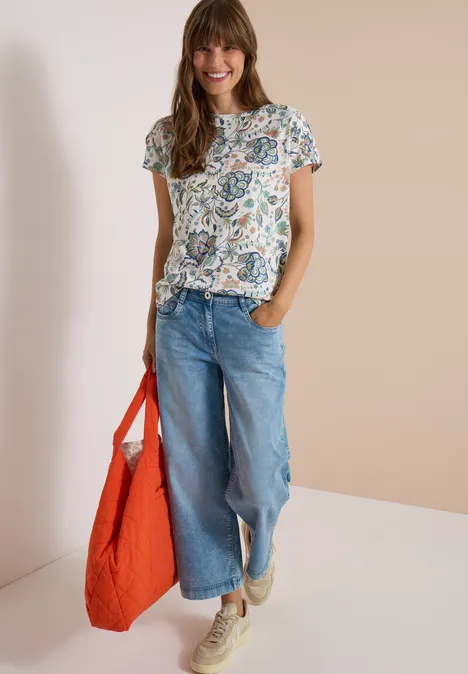 Vrouw met bloemig t-shirt, wijde spijkerbroek, beige sneakers en grote oranje tas.